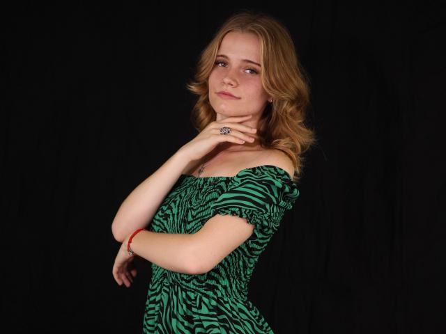 Margarita Kis, con cabello rubio y ondulado, vestido verde con estampado de cebra, con un hombro descubierto, mano derecha tocándose el cuello, fondo negro, ligera sonrisa, pulsera roja, anillo en la mano derecha..