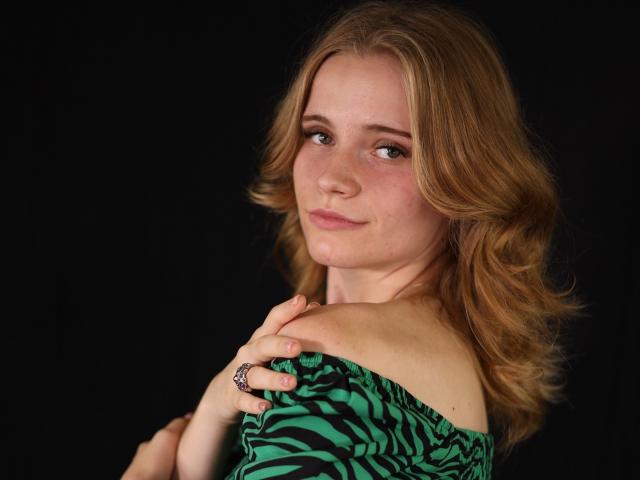 MargaritaKis with wavy blonde hair, green zebra-print dress, hands on shoulder, slight smile, dark background, ring on finger, soft lighting, cute, attractive.