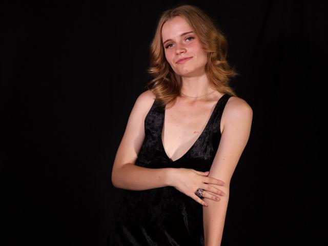 MargaritaKis with blonde hair, wearing a black velvet dress, has small tits, and a cute face, standing against a black background, smiling with one arm crossed.