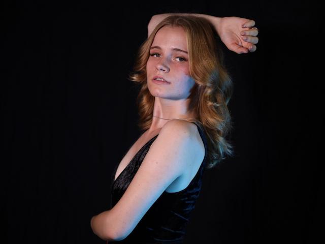 MargaritaKis with blonde wavy hair, wearing a black velvet dress, arms raised, showing her tits, pale skin, subtle smile, against a dark background, blue light on left.