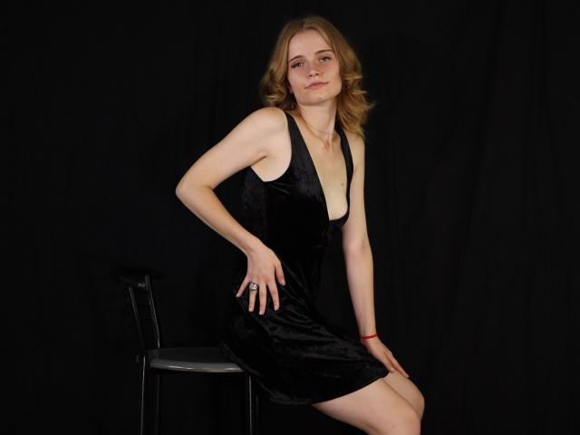 MargaritaKis with blonde hair, wearing a black velvet dress, sitting on a chair, hands on her thighs, black background, slightly leaning forward, cute, small ass, medium tits.