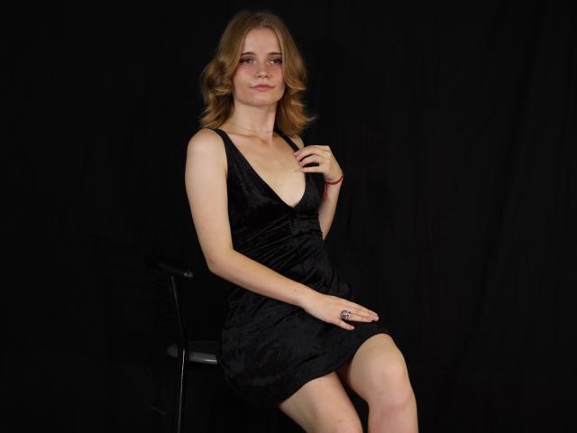 MargaritaKis with blonde hair, wearing a black velvet dress, sitting on a chair, right hand on chest, left hand on knee, black background, slight smile, medium boobs.