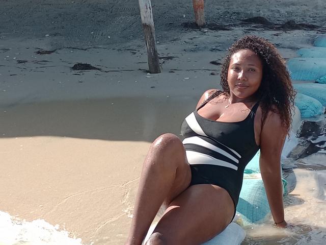 CaroldelaTorre with curly brown hair, wearing a black and white striped one-piece, sitting on a blue inflatable at the beach, showing her tits and wet ass.