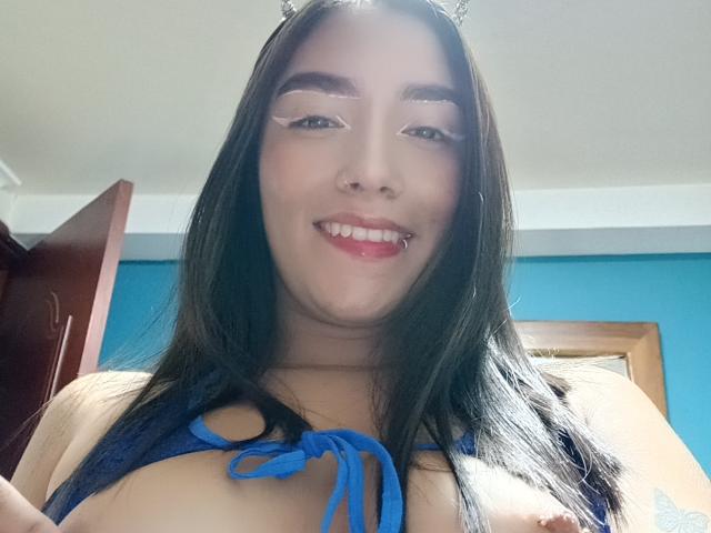 SharonKlart, con cabello negro largo, orejas de gato y un top azul de tirantes que muestra sus pechos. Tiene un piercing en la nariz, cejas gruesas y está sonriendo con un labio perforado..