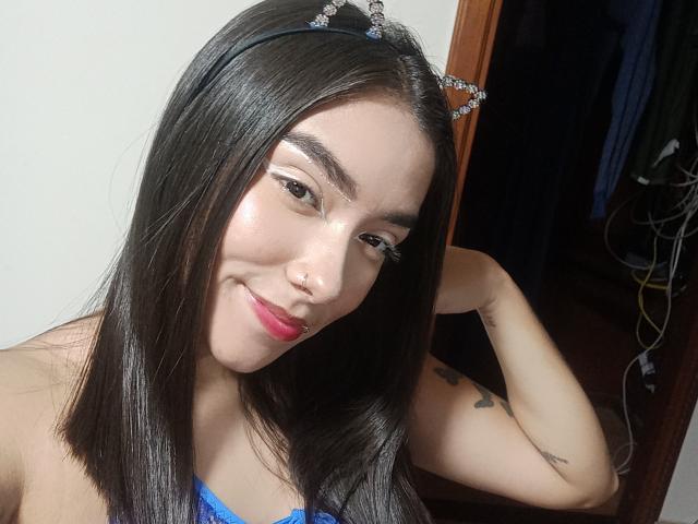SharonKlart has straight black hair, wearing a blue top, cat ears headband, and red lipstick, smiling with one hand behind her head, showing a tattoo on her arm.