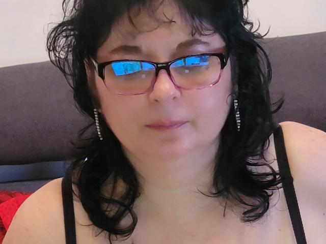 HotAndSexyMature, con capelli neri ricci, occhiali rosa e orecchini di perle, indossa una canotta nera a spalline, ha un'espressione neutra e la pelle chiara. Riflessi negli occhiali..