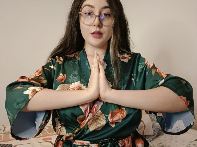 MabyLegrand with long dark hair, glasses, and red lipstick, wearing a green floral kimono, hands pressed together in a prayer position, sitting on a bed with floral sheets.