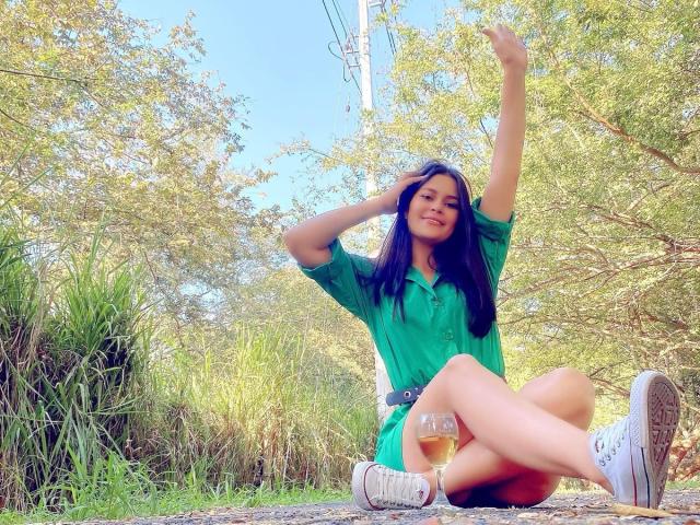SweetNaughty69 with blue hair, green shirt, and white sneakers, sitting on grass, legs crossed, arm raised, smiling, holding a glass of beer, visible pussy, trees in background.