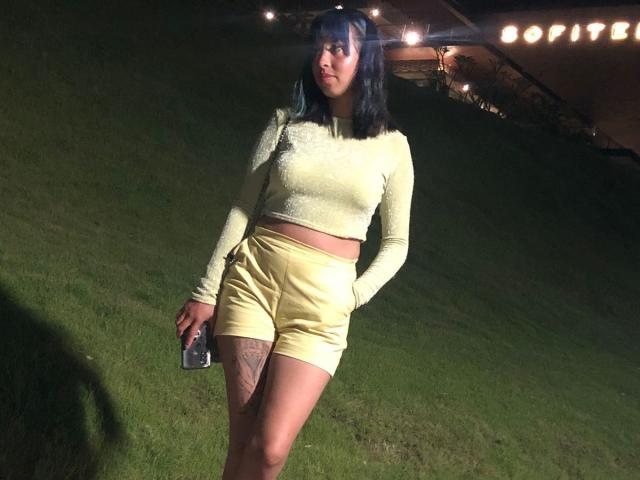 Samantha Morgan, met een bleekgeel, lang袖 crop top en gele shorts, houdt een telefoon vast, staat op gras 's nachts, heeft zwart haar, een tatoeage op haar rechter dij en kijkt naar de zijkant..