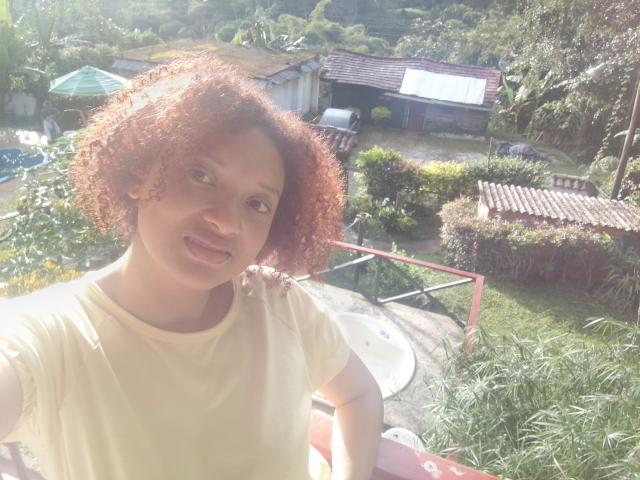 VioletaSwitch with curly red hair, wearing a yellow shirt, smiling in a sunny garden, holding a pink railing, her small tits slightly visible, green foliage in the background.