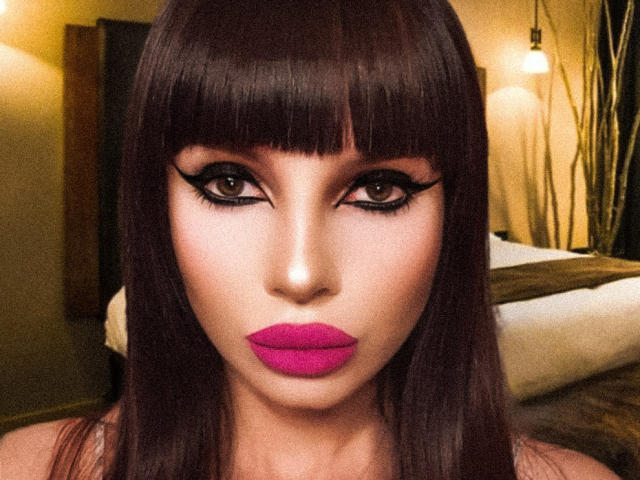 GoddessKaterina a les cheveux brun foncé, de longs cils et du rouge à lèvres rose. Ses yeux sont fixés directement sur l'appareil photo avec une expression sérieuse. Les seins ne sont pas visibles, l'attention est portée sur le visage..
