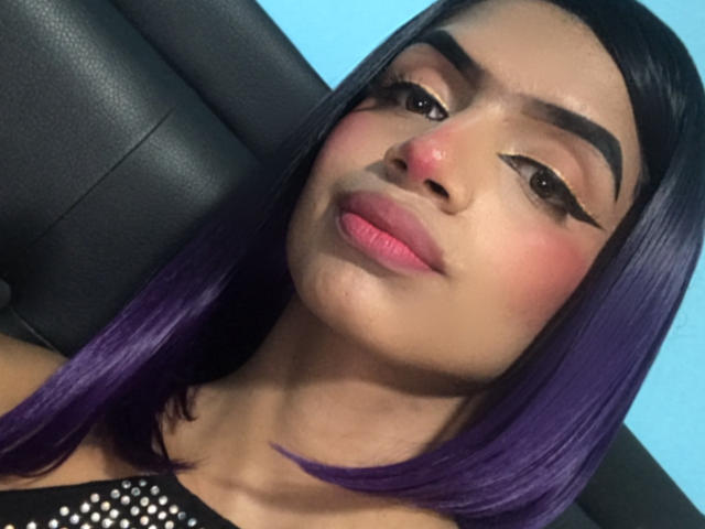 FannyBella has dark purple hair, thick eyebrows, black eyeliner wings, pink lips, and a slight smile. Her skin is tan with a rosy blush on her cheeks.