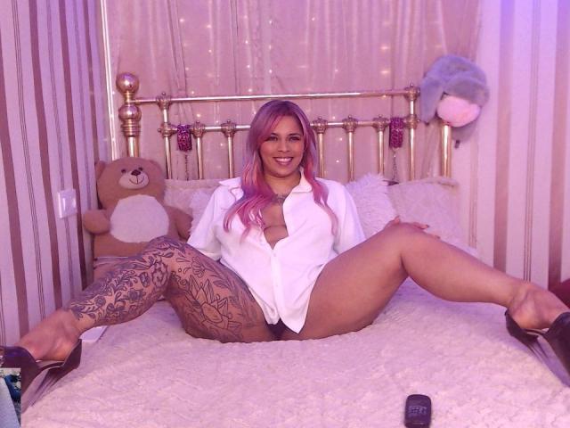 Chili con el pelo rosa, camisa blanca y medias de encaje está abriendo las piernas en una cama, sonriendo, usando tacones negros.