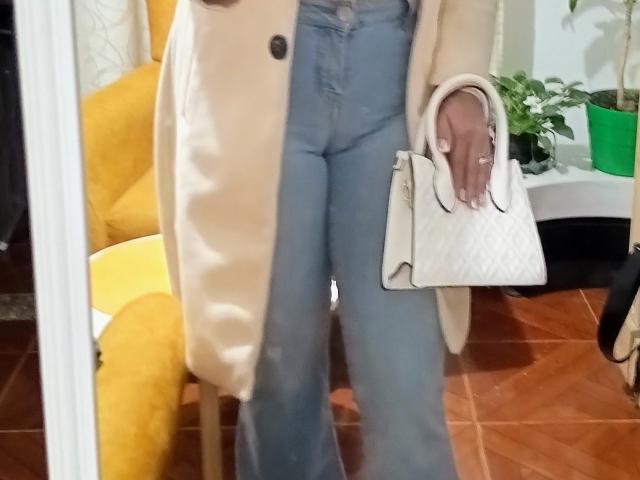 LuluFrancaise's ass is in light blue flared jeans, wearing a beige coat, holding a white quilted handbag, standing on wooden floor, yellow chair in the background.