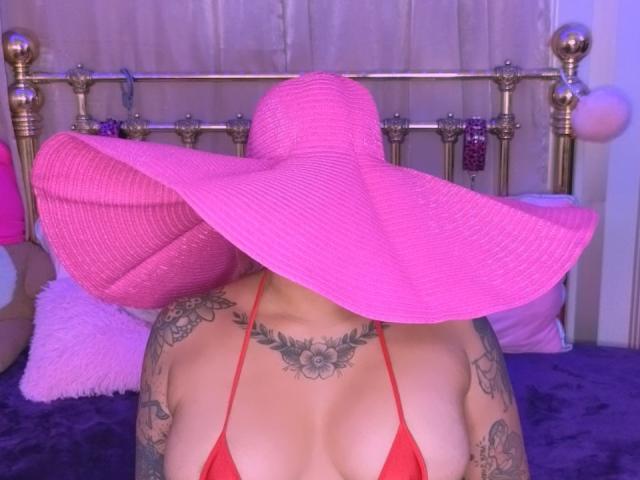 Chili's face hidden by a huge pink hat, back tattooed, wearing a red string bikini, sitting on a bed.