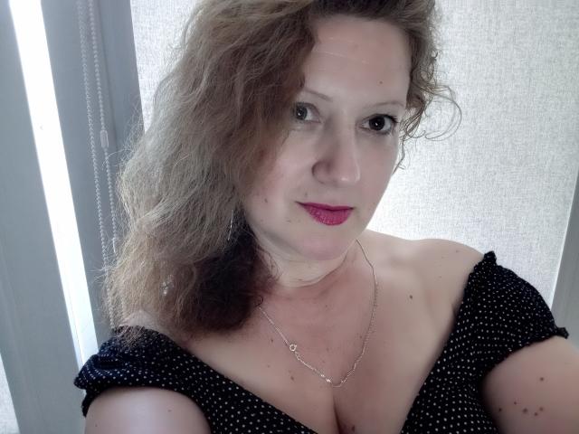 A AriaxKiss tem cabelos castanhos ondulados, pele clara e batom rosa. Ela está vestindo uma blusa preta com um ombro de fora e bolinhas brancas, que deixa seus seios à mostra. Ela tem um colar de prata..