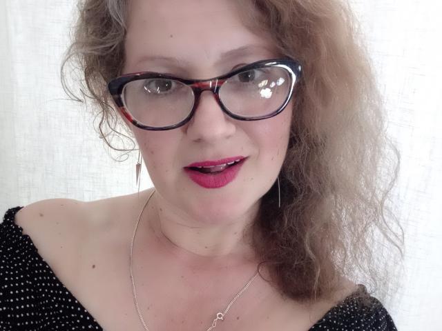 AriaxKiss ha i capelli castano ricci, indossa occhiali da vista neri a tartaruga e ha il rossetto rosso. Sta guardando la fotocamera con un leggero sorriso, indossando una maglietta nera a pois..