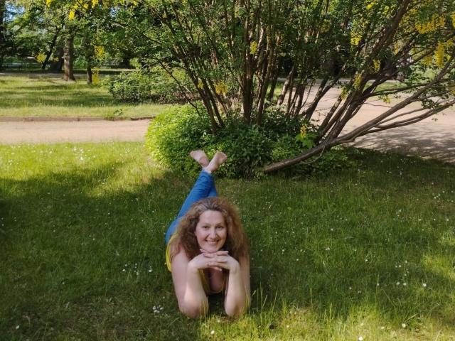 AriaxKiss è sdraiata sull'erba, con le gambe sollevate, sorridendo e con le mani sotto il mento, indossa jeans blu e una maglietta nera, piedi nudi, capelli rossi ricci, un cespuglio verde e una strada sullo sfondo..