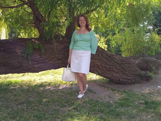 AriaxKiss, con senos de tamaño medio, vistiendo una blusa verde con un hombro descubierto y una falda blanca, sosteniendo un bolso blanco, de pie debajo de un árbol grande, sonriendo, en un área con césped..