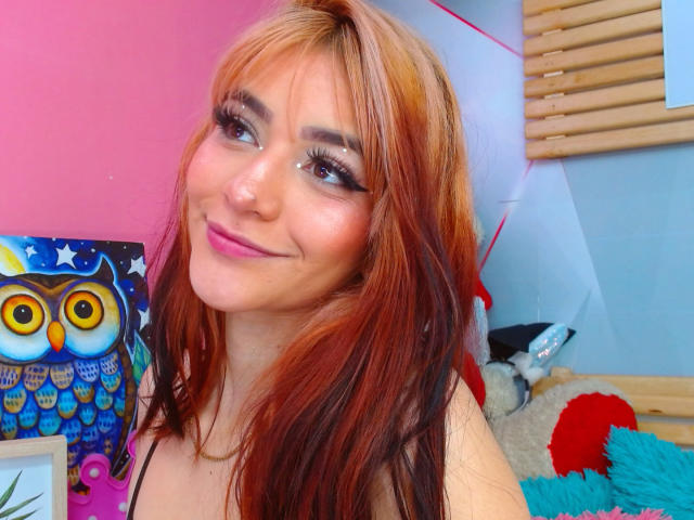 LindaWatsong with red and orange hair, smiling, wearing makeup, holding a colorful owl painting, blue eyes, pink lipstick, and long eyelashes, in a brightly colored room.