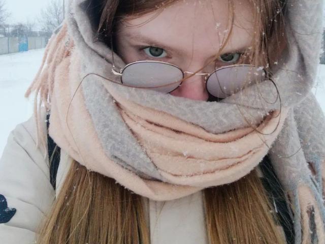 Das Gesicht von LaPochka ist größtenteils von einem dicken, rosa und grauen Schal bedeckt, wobei nur ihre grünen Augen und die rosa getönten Brillen sichtbar sind. Schneeflocken befinden sich in ihrem braunen Haar..