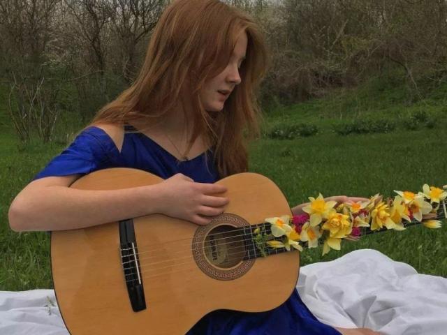 LaPochka met rood haar, een blauwe jurk, die een akoestische houten gitaar vasthoudt, versierd met gele en roze bloemen, zittend op een witte doek in een grasveld..