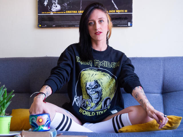 Eine Frau mit schwarzen Haaren, Tattoos und rotem Lippenstift, die ein schwarzes Iron Maiden Sweatshirt trägt, sitzt auf einem grauen Sofa, die Beine gespreizt, weiße Socken, ein gelbes Kissen und eine bunte Tasse..