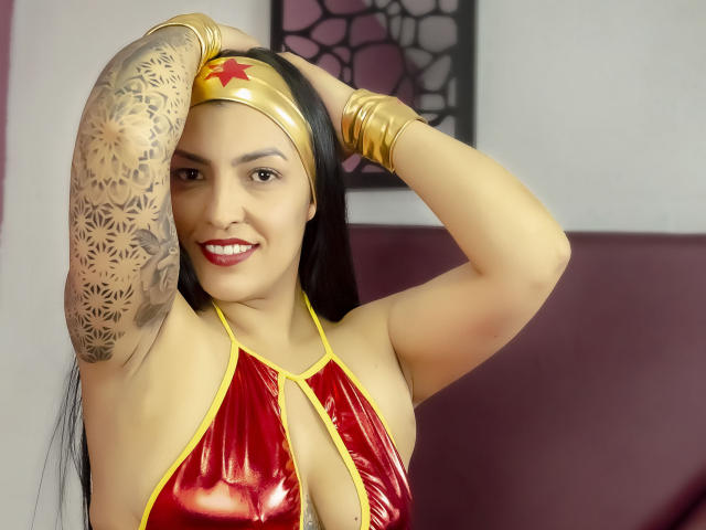 JanetteRider, con cabello negro largo, un top escotado rojo brillante, una diadema dorada, tatuajes en los brazos, sonriendo, con las manos en el cabello, pulseras doradas, mostrando sus pechos, lápiz labial rojo..