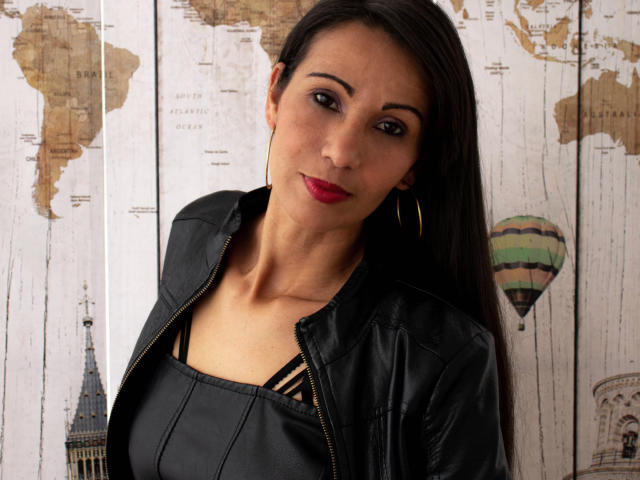 LauraFoxh, con cabello oscuro, lápiz labial rojo, chaqueta de cuero negro y aros de oro, mostrando sus senos en una blusa negra, mirando a la cámara..