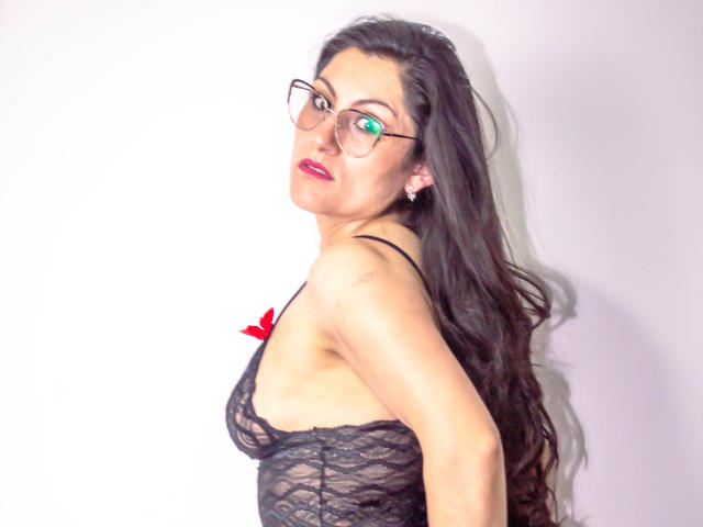 RapZelly, avec de longs cheveux noirs, des lunettes et du rouge à lèvres, portant un soutien-gorge en dentelle noire, montrant ses seins, les bras derrière le dos, fond blanc..