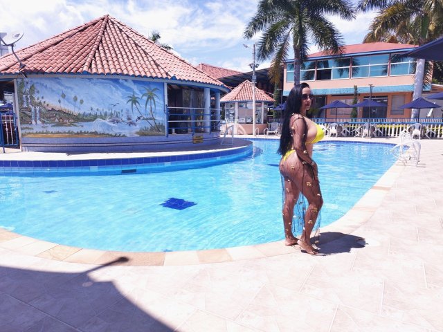 ScarletteStones con piel oscura, cabello negro largo, bikini amarillo y verde, sosteniendo un flotador amarillo, de pie junto a una piscina azul, marcas de bronceado en sus glúteos, día soleado, palmeras en el fondo..