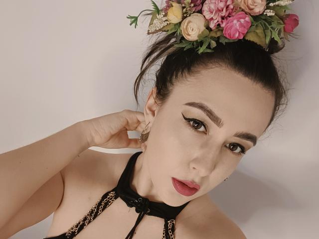 MabyLegrand with dark hair in a bun, wearing a flower crown, black halter top, and red lipstick, has prominent eyebrows and a serious expression, touching her ear.