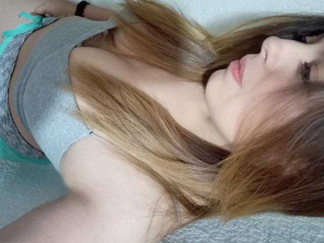 Seher with long brown hair, grey tank top, and grey shorts, showing her tits and small ass, wearing a black wristband, lying down, looking to the side.