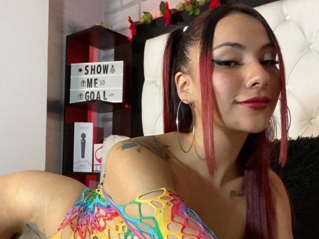 ValeryHarllow, avec des cheveux roux et noirs attachés en queue de cheval, portant un haut décontracté aux couleurs vives et à motifs arc-en-ciel, laissant apparaître ses tatouages, de grandes boucles d'oreilles et un léger sourire, regardant vers le bas..