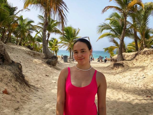 Milliee hat kleine Brüste, trägt einen einteiligen, pinken Badeanzug und eine weiße Halskette. Sie steht auf einem sandigen Strand mit Palmen, und im Hintergrund ist ein blaues Meer zu sehen..