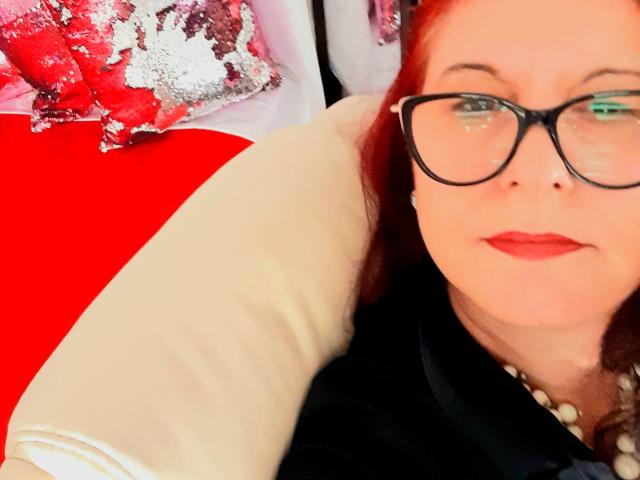 UnderMySpellK has red hair, glasses, and red lipstick, wearing a black top and pearl necklace, lying on a red and white pillow, with a white pillow behind her.