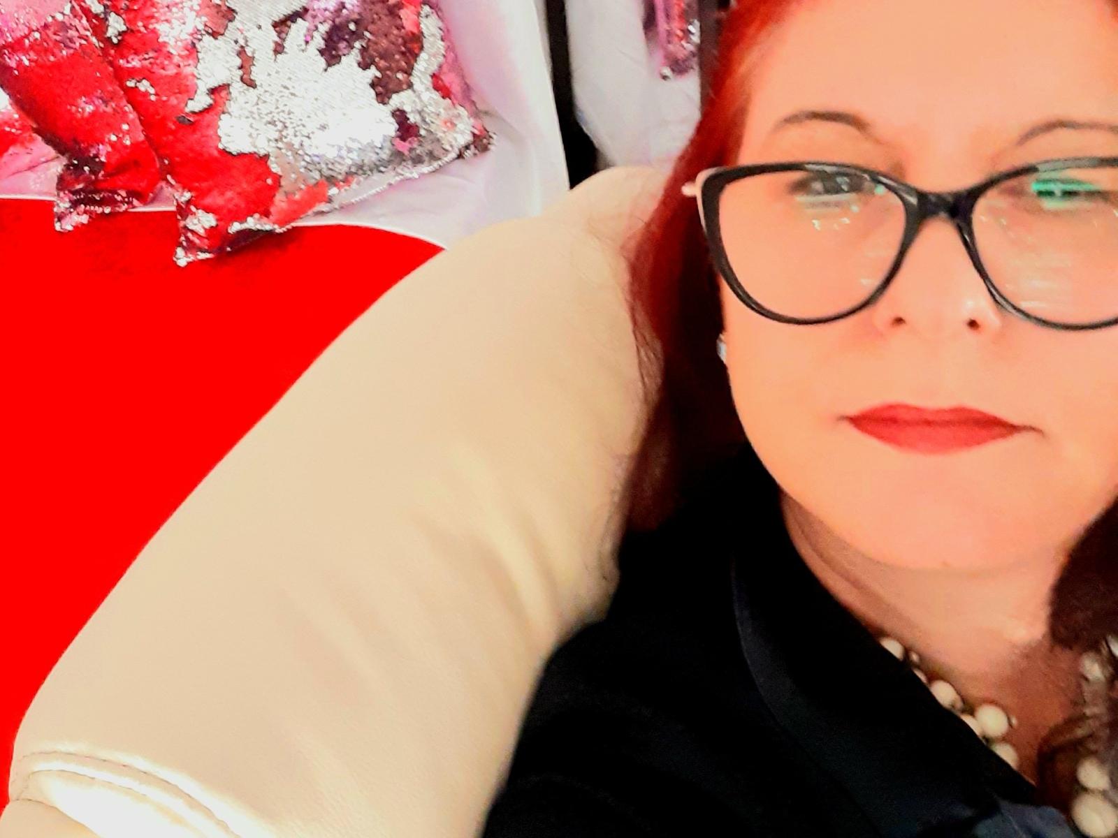 UnderMySpellK has red hair, glasses, and red lipstick, wearing a black top and pearl necklace, lying on a red and white pillow, with a white pillow behind her.