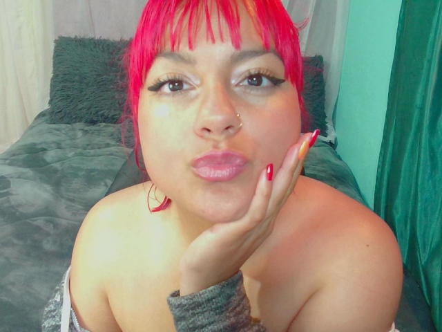 EmmaLuna, com o cabelo rosa vibrante, lábios rosados e unhas vermelhas, tocando sua bochecha com a mão, mostrando seu piercing no nariz, e usando uma manga cinza..