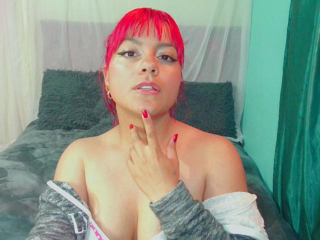 EmmaLuna mit hellroten Haaren, Nasenring und roten Nägeln, oben ohne, grauer Pullover offen, Hand auf der Brust, wobei ihre Brüste sichtbar sind, grüne Wand im Hintergrund..