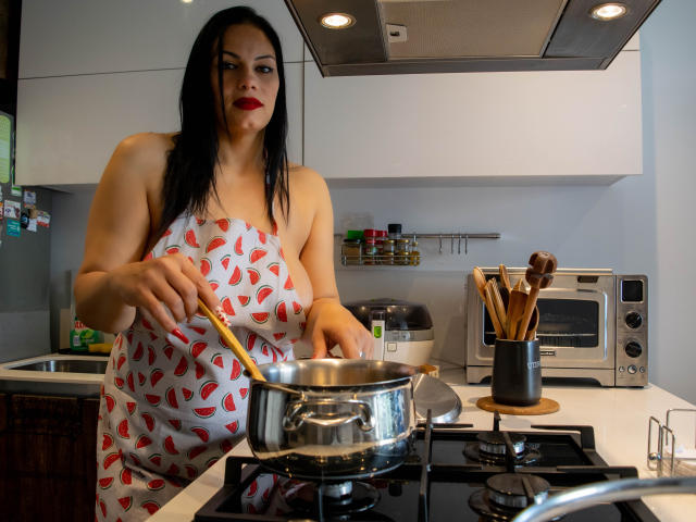 LizaRussell with black hair, red lipstick, and white apron with red cherry pattern, stirring in a stainless steel pot on a black stovetop, standing in a modern kitchen.