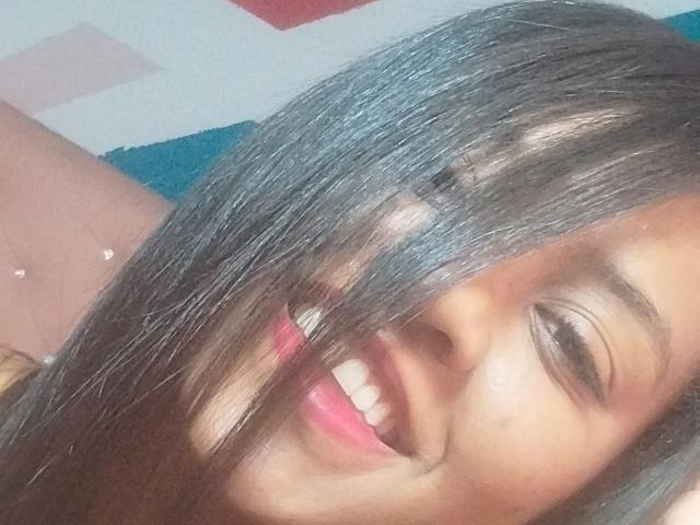 Il viso di Tiffany, con i capelli neri e lucidi che coprono un occhio, sorride con le labbra rosa, gli occhi marroni e la pelle chiara. Il suo seno è parzialmente visibile sul lato sinistro..