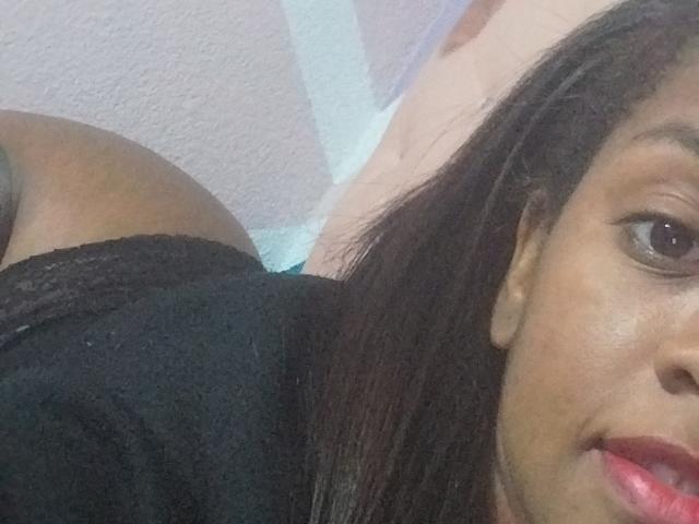 TiffanySammy with dark skin, black lace underwear, and a visible butt in the foreground. Her face, with brown eyes and red lips, is partially in the frame.