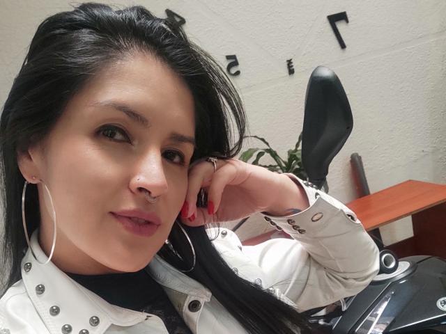Adafieldd tem cabelos pretos longos, um piercing no nariz e veste uma jaqueta branca com grandes brincos de argola prateados, unhas vermelhas e um leve sorriso, apoiada em uma cadeira preta..