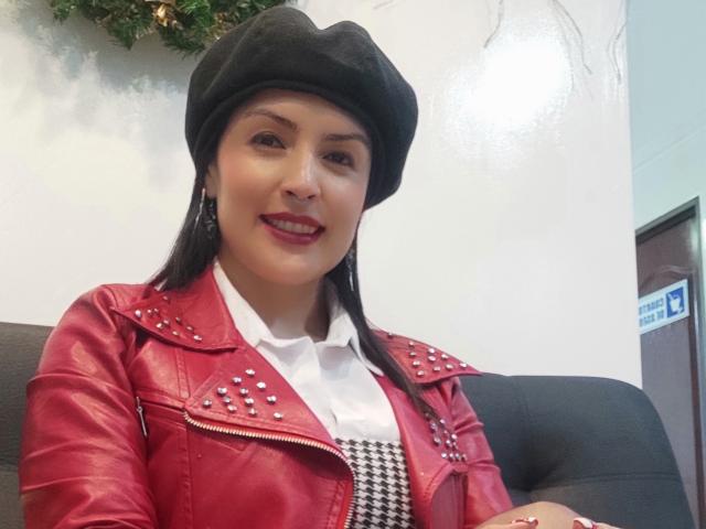Adafieldd, con senos de tamaño medio, vestida con una chaqueta de cuero roja con tachuelas, un sombrero negro, una camisa blanca y un vestido de cuadros de espiga, sonriendo a la cámara..