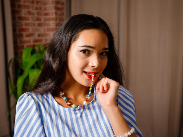 Ashley Kane, con i capelli scuri e ondulati, una camicia a righe blu, una collana di perline colorate e rossetto rosso, morde delicatamente una collana di perline, sorridendo. Ha il seno piccolo ed è carina..