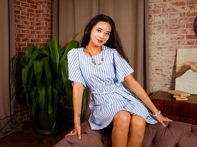 AshleyKane, with long dark hair, wearing a blue-striped dress, sitting on a brown couch, showing her ass, wearing a necklace and bracelet, smiling at the camera.