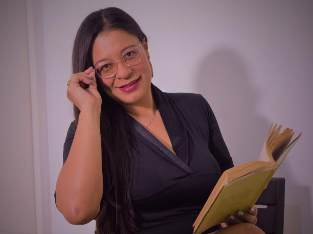 MiaCallie with long black hair, wearing glasses, a black dress, and holding a book, smiling slightly, touching her ear with her right hand. She has medium tits.