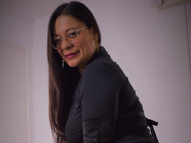 MiaCallie with long black hair, glasses, and red lipstick, wearing a black top, smiling, showing her ass, wearing gold earrings, in a slightly dim room.