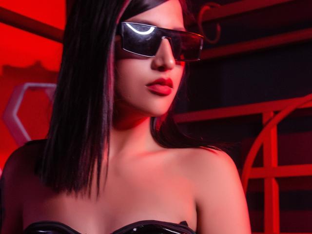 Giuliana Fetich mit schwarzen Sonnenbrillen, glattem schwarzem Haar, roter Beleuchtung, einem glänzenden, schwarzen Top ohne Träger und roten Lippen, die ihre Brüste zeigt und einen sexy, selbstbewussten Ausdruck hat..