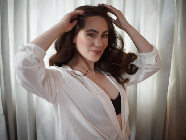 Lucia, con capelli castano ondulati, indossa una camicia bianca aperta che lascia intravedere un reggiseno nero, le mani tra i capelli, sorride, illuminazione soffusa, espressione delicata. Seno visibile, trucco leggero, posa rilassata..